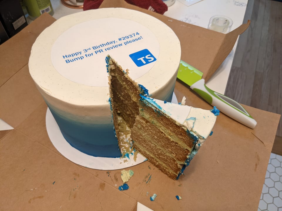 Blue and white cake with text 'Happy 3rd birthday, #29374 Bump for PR review please!' and the TypeScript logo. A tall slice is removed.