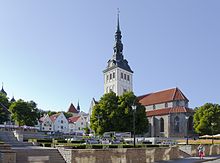 Frontale Farbfotografie von einem weißen Kirchturm mit dunkler Turmlaterne und einem Kirchenschiff im gotischen Stil. Das Kirchenschiff hat eine graue Steinfassade und ein rotes Dach.Eine Mauererhöhung mit Treppen führt zur vorderen Grünanlage.