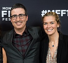 Photo of Oliver, wearing a dark suit jacket, and Norley, who is wearing a black suit jacket, standing in front of a press backdrop. Oliver has his arm around Norley.