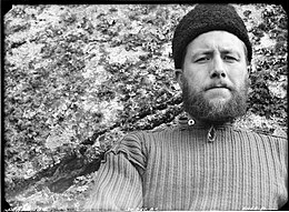 Photo noir et blanc, portrait en buste d'un homme portant la barbe et un gros bonnet sombre, devant une roche parsemée de lichens.