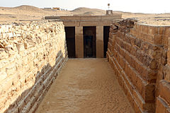 Entrance of a tomb with two columns set between long descending walls of stones.