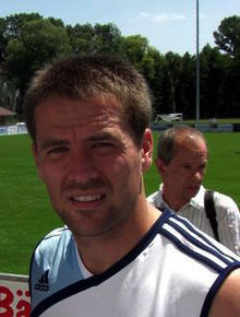 A man in a white sleeveless top, with another man in the background.