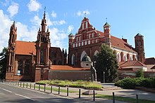 Seitliche Farbfotografie von zwei braunen Kirchen. Die linke Kirche hat zwei gotische Türme mit vielen Verzierungen. Die rechte Kirche hat vorne zwei kleine Ecktürme und ein großes, braun-weißes Giebelfeld mit einem gemalten Jesuskreuz. Hinter der Kirche ist ein breiter Turm im gotischen Stil.