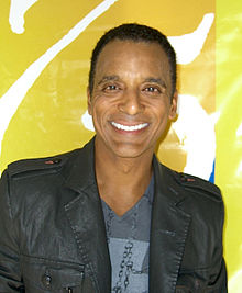 A man with short black hair, facing front, wearing a gray jacket and blue shirt.