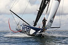 Bateau avançant sous voile, vu de devant. Le skipper est debout à l'avant. On voit la côte en arrière-plan.