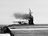 A high-wing, twin-engined aircraft taxies on the deck of an aircraft carrier, a plume of smoke rising from the carrier's island as another aircraft sits ready next to it.