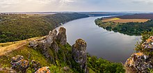 Un petit éperon rocheux, portant un drapeau, domine des reliefs de campagne basse et une large fleuve.