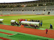 Photographie d'un stade de football, avec plusieurs joueurs sur la pelouse, alignés et de dos.