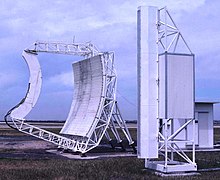 Colour photograph of two large white-painted items of equipment (the left being a parabolic 'dish' with straight sides; the right a tall, narrow structure) on grass, with cloudy sky above.