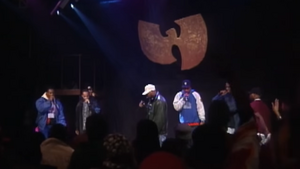 Several members of the Wu-Tang Clan performing on a stage in front of a giant logo shaped like a "W"