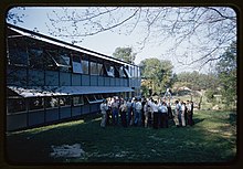 photographie d’un bâtiment à gauche et un groupement de personnes sur un espace vert au centre.