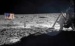 A man in a white spacesuit stands faces a spacecraft on a rocky, desolate surface; to his left stands the American flag.