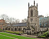 A substantial stone church seen from the northwest. The tower has crocketted pinnacles, and the north wall of the body of the church has a series of large rectangular windows