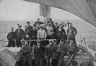 A group of men on board a ship, identified by a caption as "The Weddell Sea Party". They are dressed in various fashions, mostly with jerseys and peaked or other hats. The rough sea in the background suggests they are sailing into stormy weather.