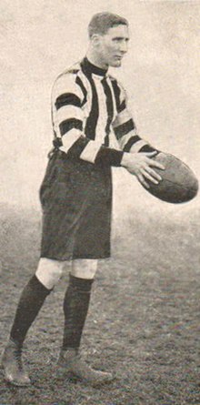 A dark-haired footballer in a long-sleeve black-and-white vertically-striped guernsey and black shorts holding a football