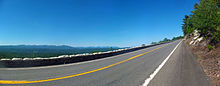 A two-lane road gently curves upward from the picture into a wooded area at right. Beyond is a line of distant mountains, green under a blue sky