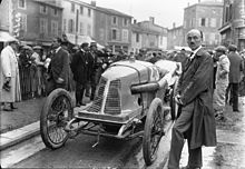 Photo d'un homme en costume posant devant une voiture, dans la rue.