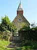 The end of a rough stone church with a tall spire-topped belfry at its far end, seen from below some steps leading to a gate