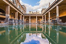 Photograph of the Baths showing a rectangular area of greenish water surrounded by yellow stone buildings with pillars. In the background is the tower of the abbey.