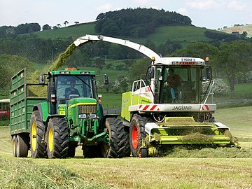 En traktor med anhænger kører ved siden af en selvkørende Claas Jaguar finsnitter.