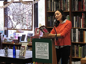 A photograph of Kerry Howley. She is standing at a podium with a Politics and Prose sign, in front of a large shelf of books. In the background is a window with a carved wooden map of the District of Columbia hanging in it. Howley has her eyes turned down, reading from her book. She is wearing an orange sweater with the sleeves pushed up and a grey collar shirt underneath.