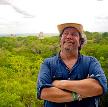 Ed Barnhart wearing a navy blue and a tan brimmed hat, standing with arms crossed, looking up toward the right with a smile on his face, Tikal in the background