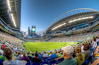 Pohled na zelený obdélníkový stadion, jež je ze všech stran obklopen tribunami plných lidí. Obě delší části tribun jsou kryty střechou, na níž jsou zavěšeny reflektory.