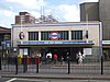 A grey-bricked building with a rectangular, dark blue sign reading "MILE END STATION" in white letters all under a light blue sky with white clouds