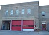 A two-story beige brick building with three garages at street level. Two have their red doors closed. Above them are metallic letters spelling out "Clay-Arsenal Station Engine Co. 2 Ladder Co. 3". On the second story are five round-arched windows together, and two more around the corner.