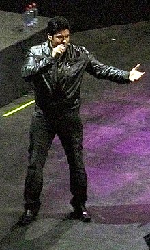 A man wearing a black jacket is performing at a stage and facing right.