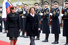 Two women in western attire walk outdoors on a red carpet along a line of soldiers in formal uniforms with rifles.