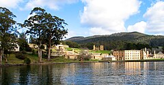 A calm body o watter is in the foregrund. The shoreline is aboot 200 metres away. Tae the left, close tae the shore, are three taw gum trees; ahint them on an incline are ruins, includin walls an watchtouers of licht-coloured stone an brick, what appear tae be the foondations o walls, an grassed auries. Tae the richt lie the ooter walls o a lairge rectangular fower-storey biggin dottit wi regularly spaced windaes. Forestit land rises gently tae a peak several kilometres back frae the shore.