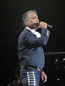 A man with grey hair is wearing a charro and holding a microphone on his right hand