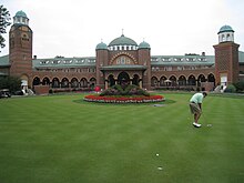 Photo du Clubhouse du Medinah Country Club avec devant un massif de fleur. Au premier plan, un joueur tente un cut.