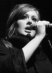 Une jeune femme, Adele, tenant un microphone dans ses mains. La photographie est en noir et blanc