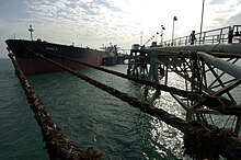 A large red and black tanker ship floats in calm dark waters, tightly moored by ropes to a long steel truss dock, while large pipes pump oil out to the ship