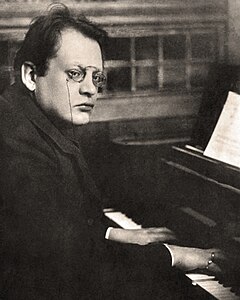 black-and-white photograph of a pianist posing at his instrument, with his hands on the keyboard at the bottom right, and his face halfway towards the camera