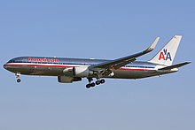 Side quarter view of twin-engine jetliner in flight, with extended gear; bare metal with red, white, and blue cheatlines, as well as two "A"s and a stylized eagle on the tail