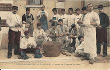 Carte postale ancienne, aux tons passés, présentant un groupe de 16 soldats en corvée de patates, dans une cour, sous la surveillance d’un officier, posant autour du baquet de bois où vont être déposés les tubercules épluchés ; les hommes, dont sept sont accroupis ou assis à même le sol, tous bottés et chapeautés d’un képi, d’un calot ou d’une coiffe, sont habillés de blanc ou portent au moins une chemise de cette couleur ; plusieurs d’entre eux sont moustachus comme l’officier ; certains sourient, d’autres font mine d’être absorbés par l’épluchage qui se fait au couteau ; au sol, les épluchures se mêlent aux légumes qui attendent d’être épluchés ; la légende dit « Les plaisirs de la caserne, Corvée de pommes de terre ».