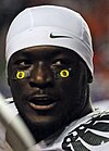 Head a young, lightly bearded, dark-skinned black man. The top of his football uniform is visible, though he is not wearing a helmet; he is wearing a white cloth cap with a Nike, Inc. logo, and two eye black patches with yellow "O"s on them. His mouth is slightly open, and his eyes are directed slightly to the camera's right.