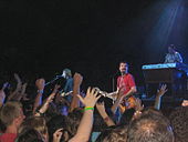 In front of a group of people with arms lifted in the air, two guitarists are playing; a keyboardist is behind them