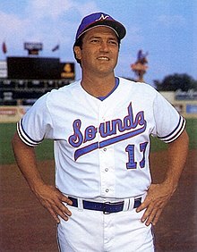 A man in a blue cap with a white "N" on the front and a white baseball uniform with "Sounds" across the chest in blue and red stands, hands on hips, on a field.