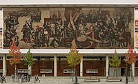 A mural consisting of red and black figures painted on concrete, depicting the history of the struggle of workers in East Germany