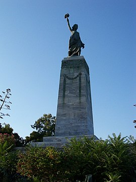 A photograph of the statue of Lerty in Mytilene; atop a podium a marble woman stands with one hand in the hair holding a torch