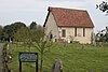 A small, red-roofed, white-walled chapel in a grassy churchyard with several saplings and gravestones