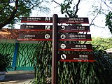 A sign labeled with directions pointing to a tourist centre, a clinic, a park exit, an animals baby farm, a peacock garden, a zoology museum, an Amusement World, and a wetland garden.