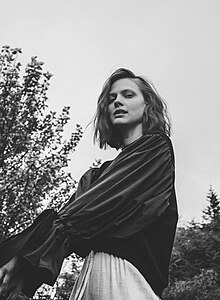 black-and-white image of Greta Svabo Bech wearing a dark top and light skirt, standing in profile, looking left at camera