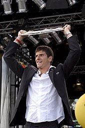 Gómez, in a suitcoat, lifts the Bundesliga trophy above his head
