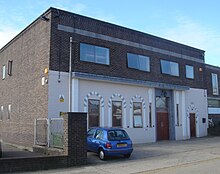 A brown-brick, flat-roofed, warehouse-style building. The lower half of its façade has been painted cream and decorated with arch-shaped window and door surrounds attached to the walls. On the first floor there are three two-pane windows and one single window. The side wall is mostly blank, with some small windows.