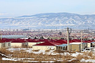 Photo d'un paysage montrant de très nombreuses maisons identiques, de plain-pied et au toit rouge, alignées selon un plan apparemment quadrillé, avec une chaîne de montagne en arrière-plan.